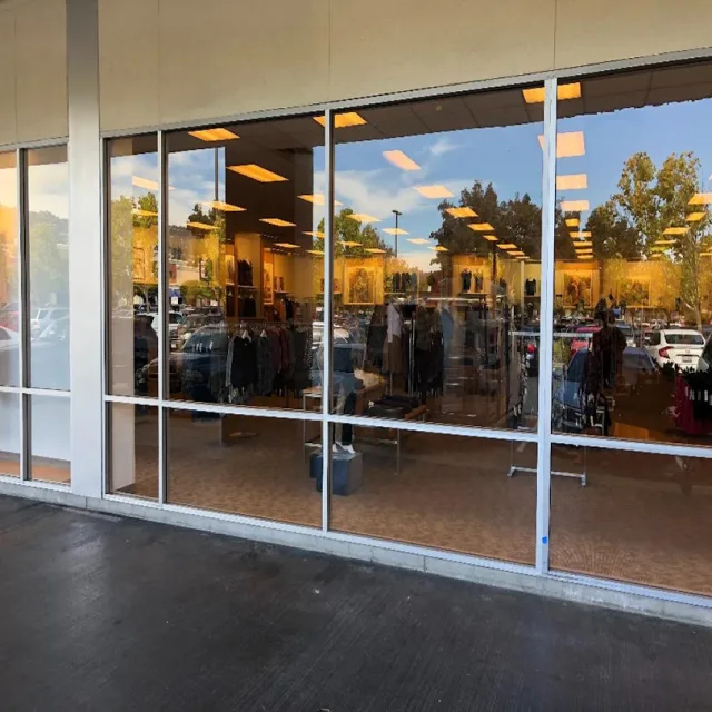 A run of tall storefront windows at a clothing store, reflecting a parking lot and trees