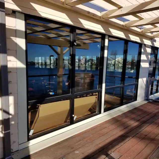 Black framed windows under a pergola reflecting open water above a wooden deck