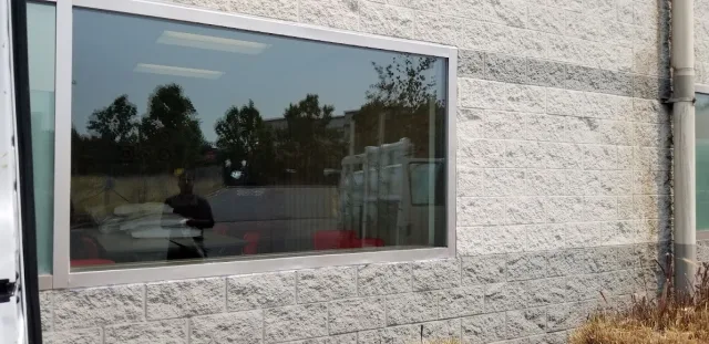 A wide fixed window in a white painted block wall, reflecting trees and a parking area