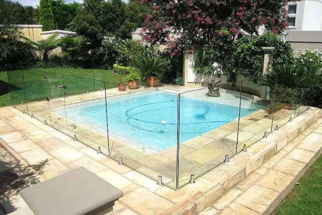 A square pool enclosed by frameless glass panels on sandstone paving in a green garden