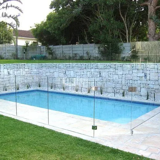 A frameless glass pool fence held in spigots around a pool, in front of a stone retaining wall