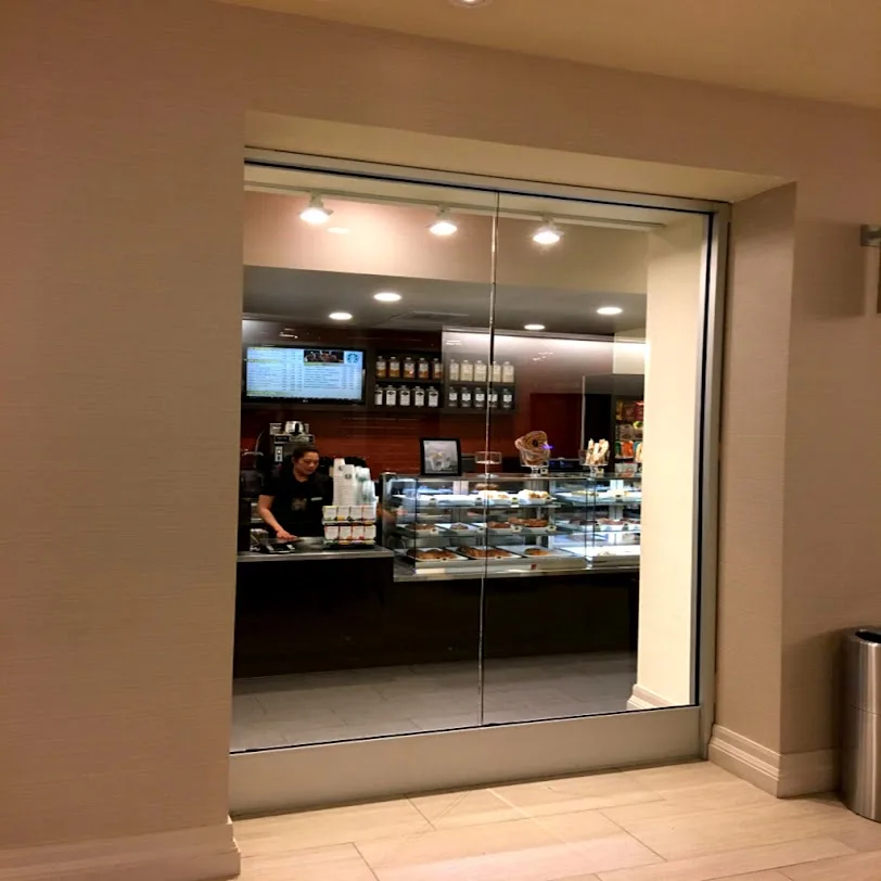 A frameless glass wall and opening looking into a hotel cafe with a pastry counter