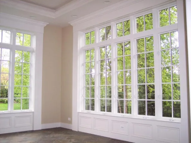 An empty room with a wall of tall white multi pane windows looking out at green trees