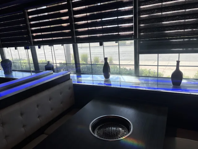 A restaurant booth with a tabletop grill beside a long run of windows with shades pulled halfway