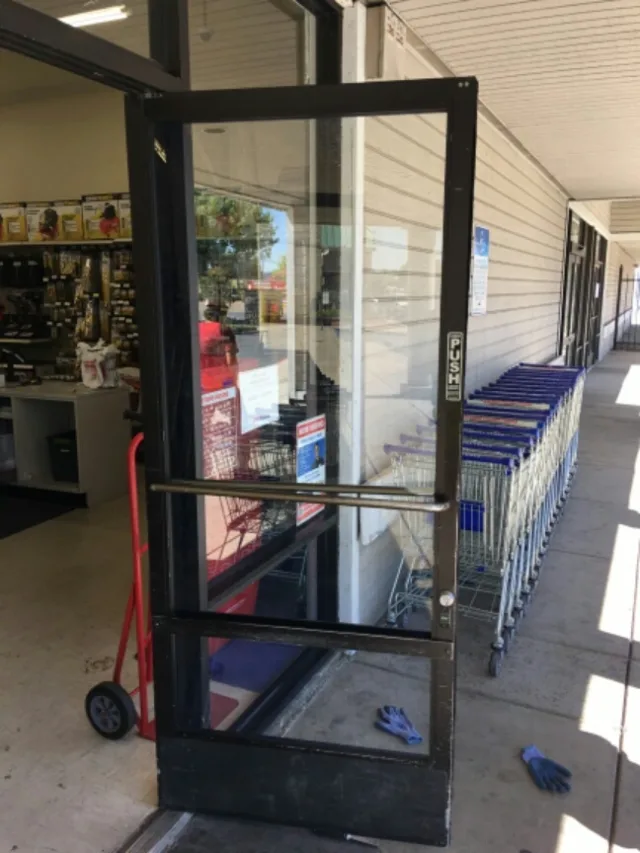A black aluminum storefront door propped open at a shop entrance, with work gloves on the walkway