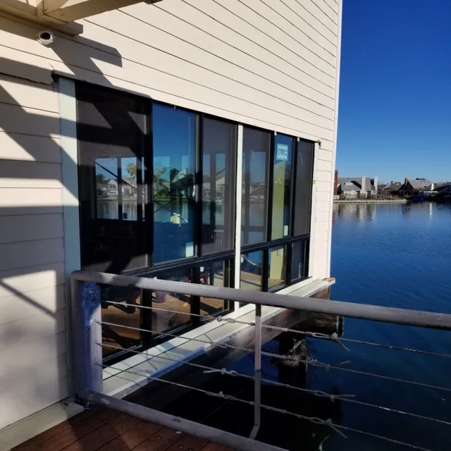 Tall dark framed windows on a light sided building above the water, with a cable railing in front